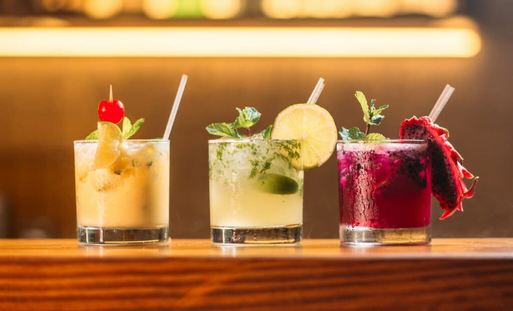 Três copos de vidro com drinks diferentes em um balcão de madeira