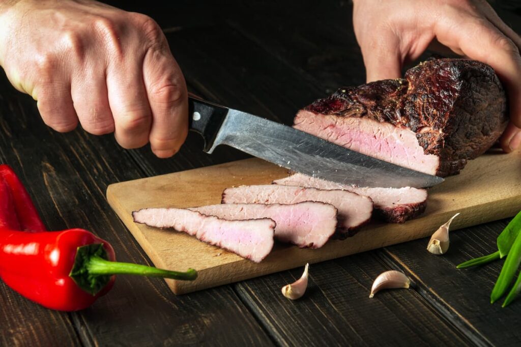 Uma pessoa cortando um pedaço de carne bem passada, em uma tábua de madeira
