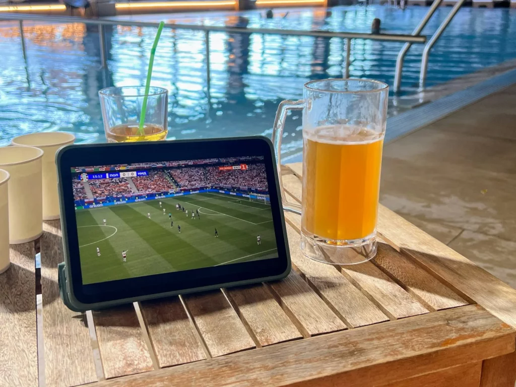 Foto de um tablet passando jogo de futebol com um copo de cerveja ao lado.